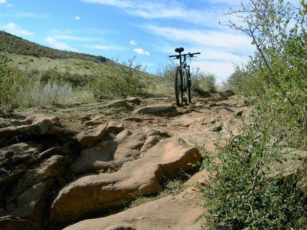 A mountain bike stands beside a rocky trail surrounded by sparse vegetation and rolling hills under a partly cloudy sky. Devil's Backbone mountain bike trail.
