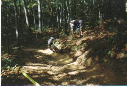 A mountain biker navigating a dirt trail in a wooded area, with trees lining the path. In the background, two people are observing the biker. The scene captures the excitement of outdoor cycling in nature. Fontana Village mountain bike trail.