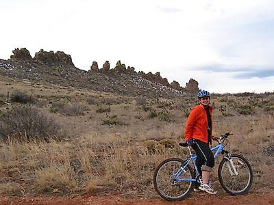 A person in an orange jacket and blue bicycle helmet sits on a mountain bike in a gravel path, surrounded by a dry landscape featuring sparse vegetation and rocky formations in the background. Devil's Backbone mountain bike trail.