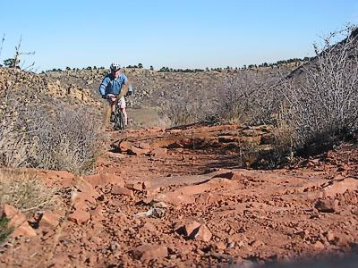 A cyclist navigating a rocky trail in a rugged landscape, surrounded by sparse vegetation and distant hills under a clear blue sky. Devil's Backbone mountain bike trail.