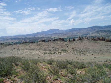A panoramic landscape view showcasing rocky formations and rolling hills under a partly cloudy sky. In the foreground, there is sparse vegetation and rocky terrain, while the background features distant mountains. Devil's Backbone mountain bike trail.