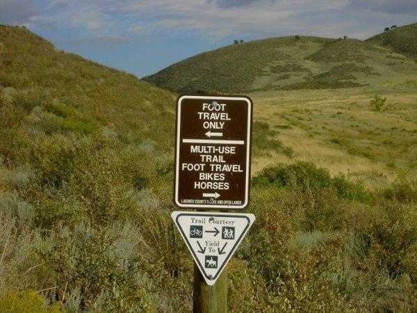 A brown informational sign in a natural landscape indicating trail usage regulations, with sections for "Foot Travel Only" and "Multi-Use Trail" for foot travel, bikes, and horses. Below, a triangular sign illustrates right-of-way rules for different trail users. The background features rolling hills and greenery. Devil's Backbone mountain bike trail.