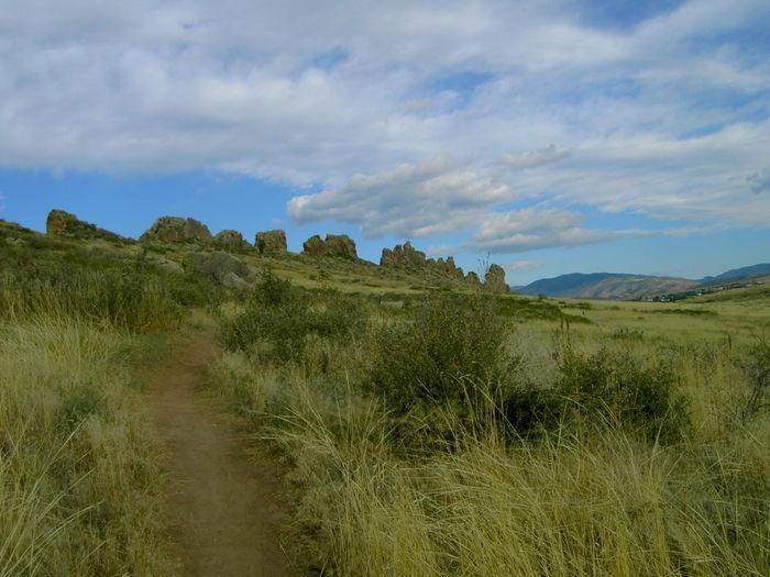 A dirt path winding through a grassy landscape, leading towards rocky formations in the distance under a partly cloudy blue sky. Devil's Backbone mountain bike trail.