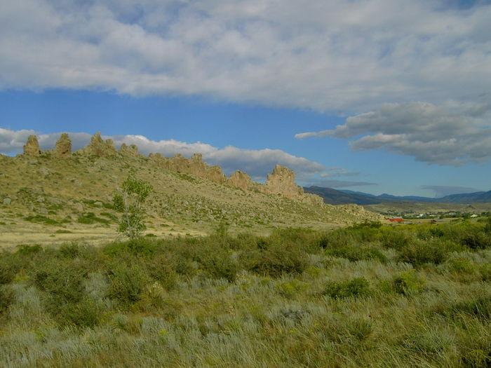 A scenic landscape featuring rugged rock formations atop a grassy hill, with shrubs and trees in the foreground. The sky is partly cloudy with hints of blue peeking through. In the distance, rolling hills and a small agricultural area can be seen, suggesting a natural and serene environment. Devil's Backbone mountain bike trail.
