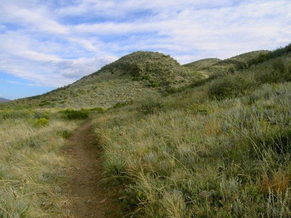 A dirt path winding through grassy hills under a partly cloudy sky. The landscape features rolling green mounds with sparse vegetation, inviting exploration of the natural scenery. Devil's Backbone mountain bike trail.