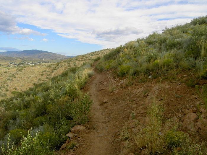 A winding dirt trail leads up a hillside covered with green vegetation. The landscape is dotted with small rocks, and in the distance, rolling hills and a vibrant sky filled with clouds can be seen. The scene captures a serene and natural outdoor setting, ideal for hiking. Devil's Backbone mountain bike trail.