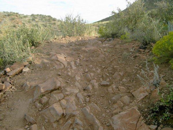 Rocky hiking trail surrounded by sparse vegetation and hills under a partly cloudy sky. The path is uneven and consists of loose rocks and dirt, suggesting a challenging terrain for hikers. Devil's Backbone mountain bike trail.