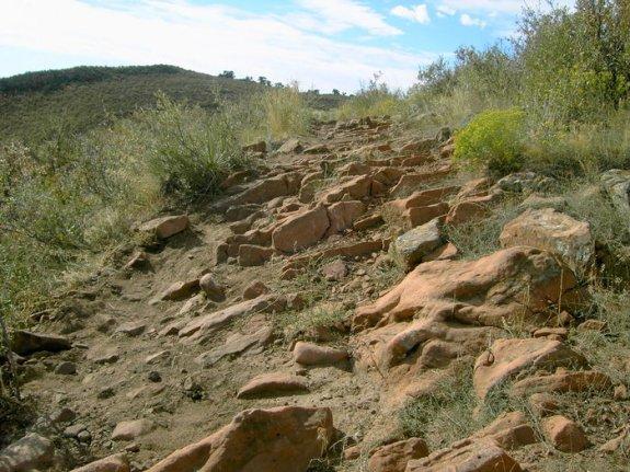 Rocky hiking path winding through a natural landscape, surrounded by sparse vegetation and hills under a bright blue sky with clouds. Devil's Backbone mountain bike trail.