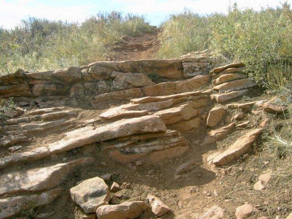 A rocky trail leading uphill, featuring uneven stone steps surrounded by sparse vegetation and dry earth. The pathway is lined with various sizes of flat and jagged rocks, with some grass and shrubs growing along the edges. Devil's Backbone mountain bike trail.