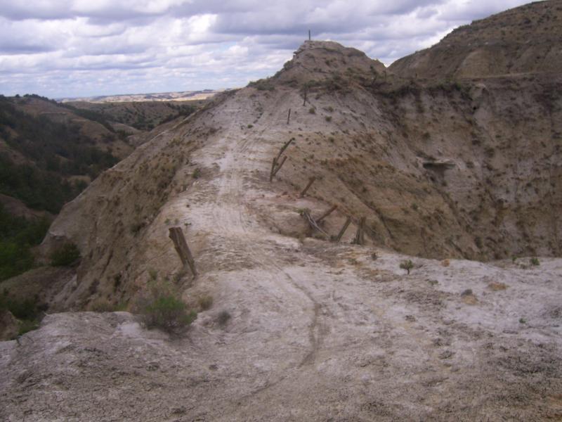 A rocky landscape featuring a narrow, winding pathway cutting through a dry, hilly terrain. The scene includes weathered wooden posts and sparse vegetation, with a cloudy sky overhead. The hills display muted earth tones and a rugged, uneven surface. Maah Daah Hey mountain bike trail.