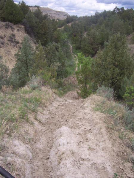 A sandy trail winding through a green, wooded area, with hills on either side and a cloudy sky above. The path is surrounded by shrubs and trees, indicating a natural landscape ideal for hiking and exploration. Maah Daah Hey mountain bike trail.
