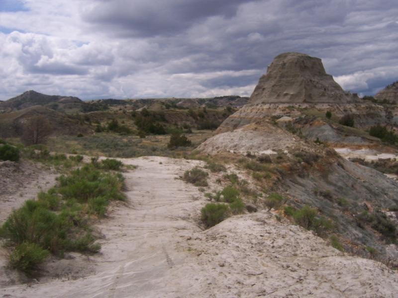 A scenic view of a rugged landscape featuring a dirt path winding through dry terrain, with unique rock formations and patches of greenery under a cloudy sky. Maah Daah Hey mountain bike trail.