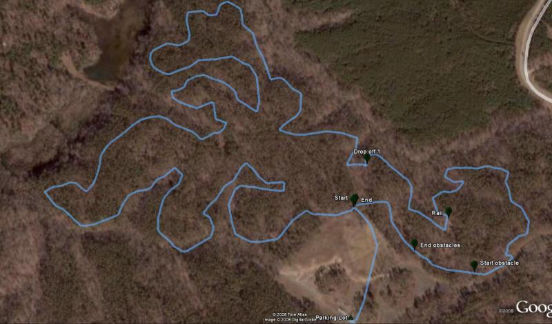 Map of a wooded area with a marked trail outlined in blue. The trail features key points labeled as "Start," "End," "Drop off 1," "Start obstacle," "End obstacles," and "Rail." The surrounding terrain is mostly bare with patches of greenery, indicating a natural landscape. The map also includes a parking lot icon near the bottom. Freedom Park (aka:district Park) mountain bike trail.