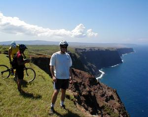 Three cyclists are enjoying a scenic view from a cliff overlooking the ocean on a sunny day. Lush green grass surrounds them, and the coastline features rugged cliffs and clear blue water. One cyclist stands in the foreground, while the others are slightly behind, all wearing helmets. Waikolu Overlook, Molokai Ranch mountain bike trail.