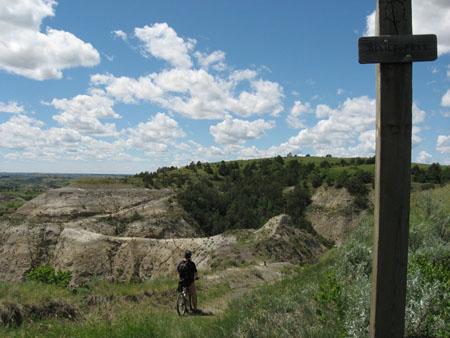 A cyclist standing on a dirt path next to a wooden signpost, surrounded by rugged hills and a blue sky with fluffy clouds. The landscape features green vegetation and rocky terrain. Maah Daah Hey mountain bike trail.