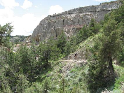 A scenic view of a rugged landscape featuring a rocky cliff with layered textures, surrounded by greenery including trees and shrubs. In the foreground, a narrow path winds through the foliage, and a person can be seen biking along the trail. The sky is partly cloudy, adding to the vibrant natural setting. Maah Daah Hey mountain bike trail.