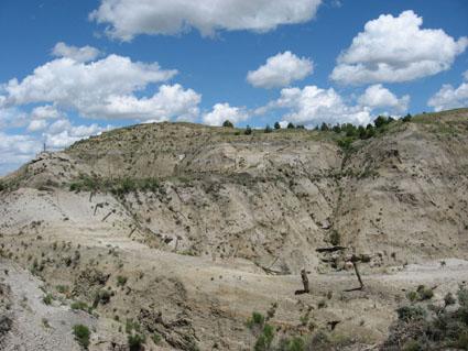 A dry, rugged landscape featuring a steep hill and rocky terrain under a bright blue sky adorned with fluffy white clouds. The surface shows signs of erosion with sparse vegetation scattered throughout. Maah Daah Hey mountain bike trail.