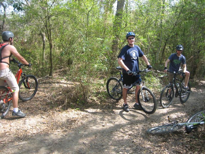 A group of three cyclists enjoying a break on a dirt trail surrounded by greenery. Two riders are standing by their bikes, while one is seated on a bike. They are wearing helmets and casual clothing suitable for biking. Sunny day with dappled light filtering through the trees. Comite Trails mountain bike trail.