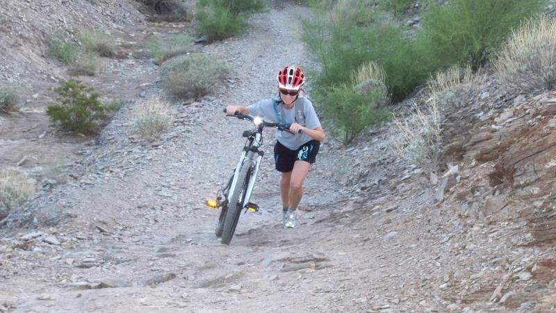 A person in athletic gear pushes a mountain bike up a rocky, uneven trail surrounded by sparse vegetation. The individual, wearing a helmet and sunglasses, appears focused and determined as they navigate the challenging terrain. Trail #100 mountain bike trail.