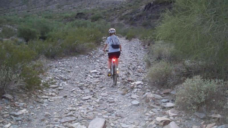 A person riding a mountain bike along a rocky trail surrounded by sparse vegetation and hills. The cyclist is wearing a helmet and cycling gear, with a red light attached to the back of the bike, indicating low light conditions. The scenery is rugged, featuring a mix of stones and desert plants. Trail #100 mountain bike trail.