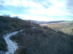 A scenic view of a winding trail through hilly terrain, surrounded by sparse vegetation and patches of snow, with mountains in the background under a partly cloudy sky. Bear Creek Lake Park mountain bike trail.