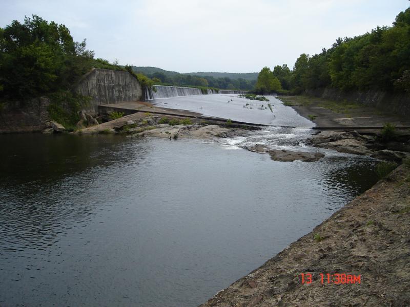 A serene view of a river with a dam in the background. The water flows over the dam, creating a waterfall effect. Surrounding the river are rocky and earthy areas with sparse vegetation, while lush green trees line the banks in the distance under a cloudy sky. Oklahoma "ankle Express" Hiking Trail - Greenleaf State Park mountain bike trail.
