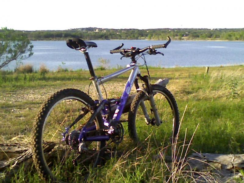 A mountain bike leaning against a log with a scenic view of a lake and hills in the background. The bike features a silver frame with purple accents, surrounded by green grass and sparse shrubs. The sky is clear, indicating a sunny day. Madrone Trail mountain bike trail.