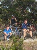 Three individuals are sitting with bicycles in a natural setting. Two are seated on the ground, while one is standing behind them. All are casually dressed, and there are trees and greenery in the background. Madrone Trail mountain bike trail.