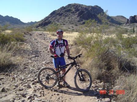 A cyclist wearing a colorful jersey and helmet stands beside a mountain bike on a rocky trail in a desert landscape, with mountains in the background and sparse vegetation around. The date displayed in the corner is November 17, 2003. Trail #100 mountain bike trail.