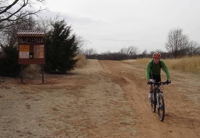 A cyclist in a green jacket and orange helmet rides on a dirt path alongside a trailhead sign that reads "Blue Creek." The background features sparse trees and grass under a cloudy sky. Bluff Creek Trail mountain bike trail.