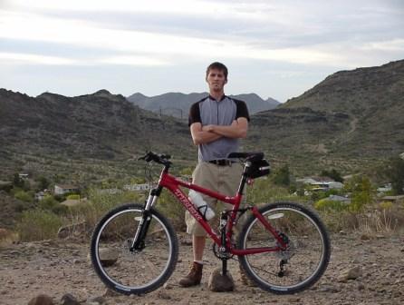A person standing confidently with arms crossed next to a red mountain bike, set against a backdrop of rocky hills and a cloudy sky. Trail #100 mountain bike trail.