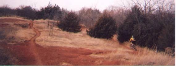 A scenic dirt trail through a grassy area with a reddish-brown soil surface, bordered by trees and shrubs. A cyclist in a yellow shirt rides along the trail on the right side of the image. The landscape features a mix of sparse vegetation and open space, with a cloudy sky overhead. Bluff Creek Trail mountain bike trail.