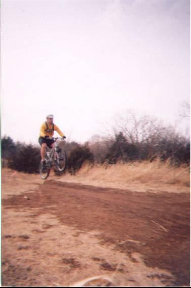 A mountain biker wearing a yellow jersey and helmet performs a jump on a dirt trail, surrounded by dry grass and sparse trees under a cloudy sky. Bluff Creek Trail mountain bike trail.