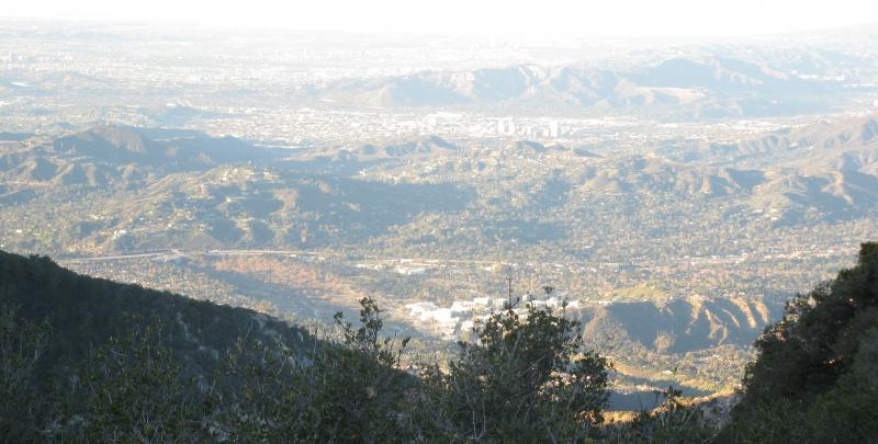 A panoramic view of rolling hills and valleys, with a cityscape visible in the distance under a clear sky. The foreground features greenery, while the landscape stretches out toward the horizon, showcasing a mix of urban and natural scenery. Gabrielino Trail mountain bike trail.