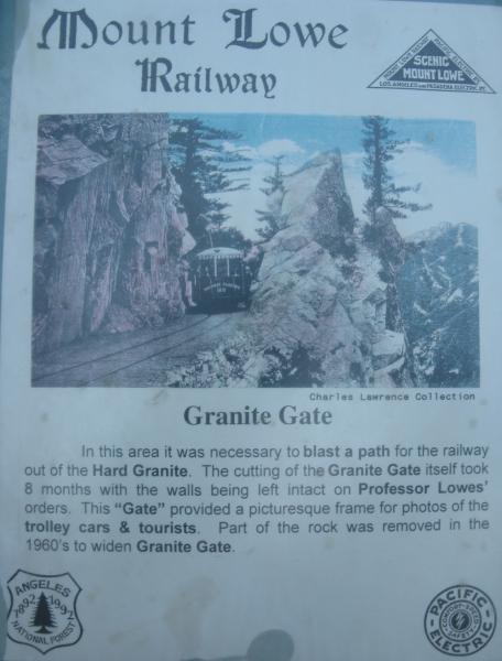 An historical photograph depicting the Granite Gate of the Mount Lowe Railway, showcasing a railway path carved through rocky terrain. The image features a train passing through the granite gateway, surrounded by tall trees and mountainous scenery. The text provides context about the construction of the Granite Gate and its significance for the railway and tourists. Gabrielino Trail mountain bike trail.