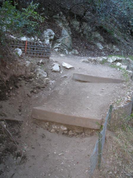 A dirt pathway leading through a wooded area, featuring two wooden steps that create a transition between levels of the trail. Nearby, there are rocks and a small fence made of wire. The surroundings consist of trees and rocky terrain. Gabrielino Trail mountain bike trail.