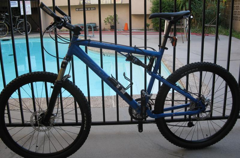 A blue mountain bike is leaning against a black metal fence, with a swimming pool visible in the background. The bike features thick tires and a suspension system, positioned on a paved surface near the pool area. Gabrielino Trail mountain bike trail.
