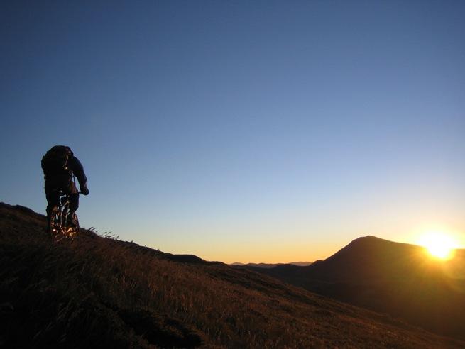 A mountain biker ascends a grassy hillside during sunset, with a vibrant orange sun peeking over the horizon and mountains in the background. The sky transitions from blue to warm hues, creating a serene atmosphere. Sunrise mountain bike trail.
