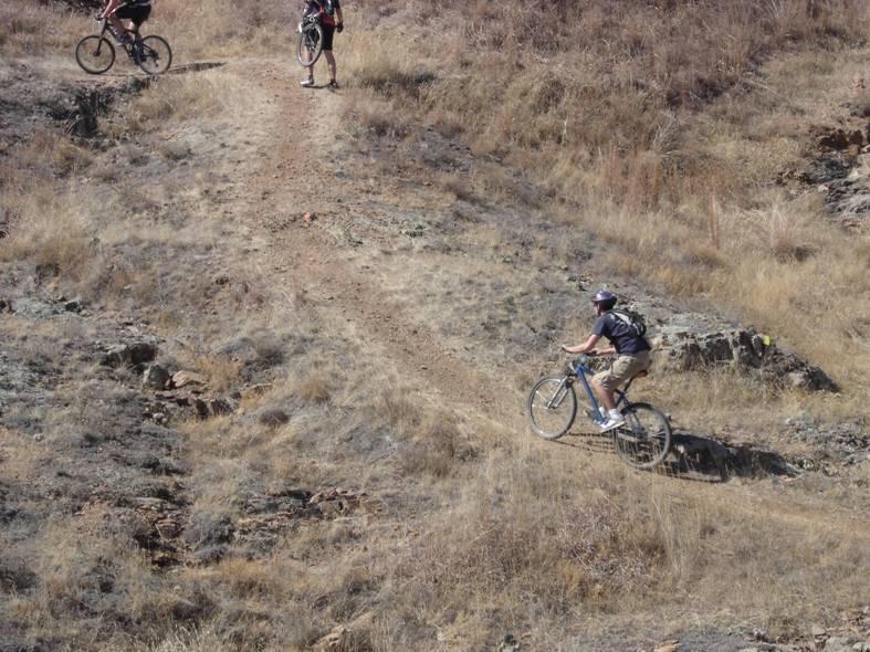 Three mountain bikers navigate a rugged trail on a dry, rocky hillside covered with sparse grass. One biker is in the foreground, riding downhill, while the other two are positioned higher up on the trail. The setting suggests a sunny day with a natural outdoor environment. 12 Miles Of Hell mountain bike trail.