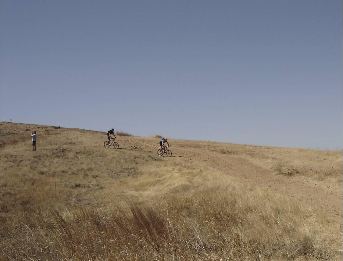 Two mountain bikers ride on a dirt trail through a dry, grassy landscape under a clear blue sky, while a third person stands nearby, observing them. 12 Miles Of Hell mountain bike trail.
