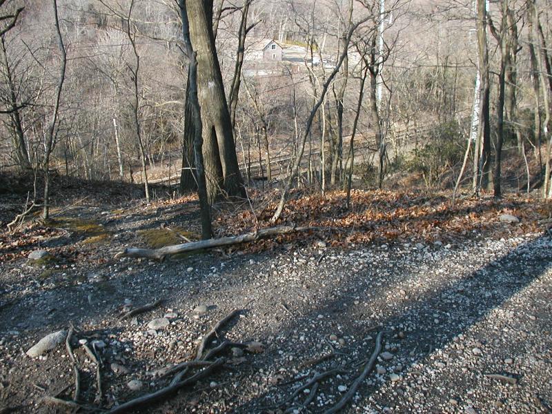 A wooded trail with a rocky, uneven surface leads downhill, surrounded by bare trees and scattered leaves. In the background, a glimpse of a small house and a fence can be seen through the branches, hinting at a nearby clearing. The scene captures a peaceful, rustic landscape in a late autumn or early winter setting. Trail View State Park mountain bike trail.