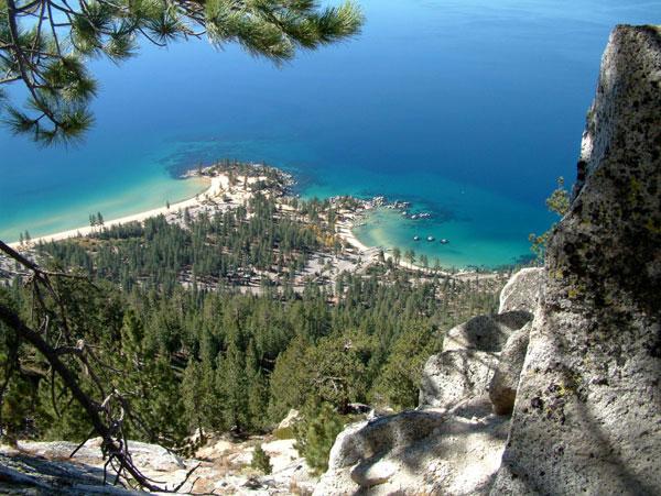 Aerial view of a scenic shoreline with sandy beaches and clear blue water, surrounded by dense green forests and rocky cliffs. The image captures a tranquil bay with boats anchored near the beach, illustrating the natural beauty of the landscape. Tahoe Rim Trail mountain bike trail.