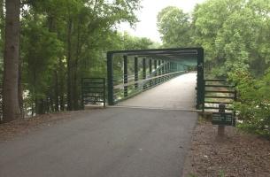 A green pedestrian bridge extends across a path surrounded by lush trees. The scene is misty, giving a tranquil atmosphere. The bridge is framed by vertical railings, leading to a pathway that disappears into the distance. North Augusta Greeneway mountain bike trail.