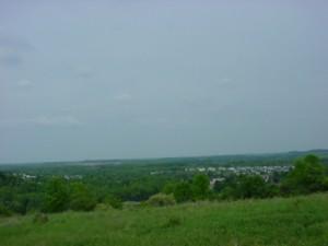 A panoramic view of a green landscape under a cloudy sky, featuring rolling hills and a distant town in the background. Indian Hill mountain bike trail.