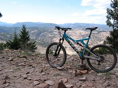 A blue mountain bike resting on rocky terrain with a scenic mountain landscape in the background under a partly cloudy sky. Heil Valley Ranch mountain bike trail.