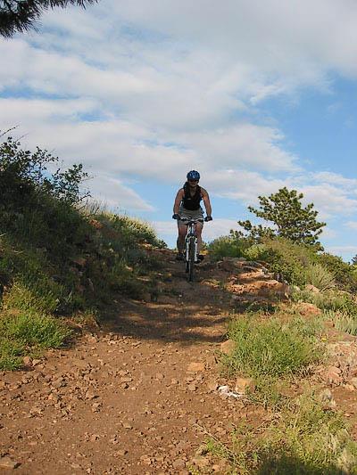 A mountain biker riding down a rocky trail surrounded by lush greenery and blue skies. The cyclist is wearing a helmet and athletic clothing. Rabbit Mountain Loop mountain bike trail.