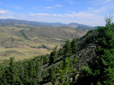 A panoramic view of rolling hills and valleys, with a foreground of evergreen trees. The landscape features a variety of green hues, rocky outcrops, and distant mountains under a clear blue sky with a few scattered clouds. Heil Valley Ranch mountain bike trail.