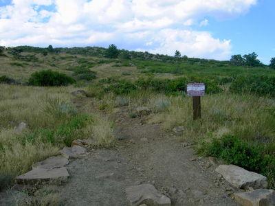 A dirt trail leading into a grassy landscape, flanked by patches of vegetation. A signpost stands beside the trail, indicating directions or information about the area. The sky is partly cloudy with scattered clouds. Rabbit Mountain Loop mountain bike trail.