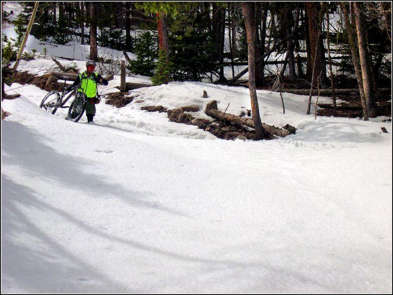 A person in a bright green jacket is walking through snow while carrying a mountain bike. The surrounding area features trees and a mixture of snow and exposed ground, suggesting early spring conditions in a forested environment. Ceran Saint Vrain Loop mountain bike trail.