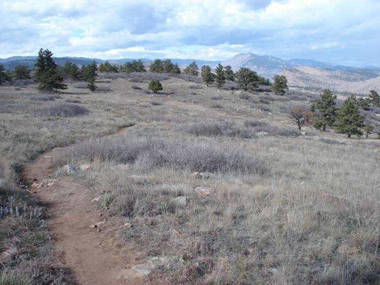 A dirt hiking trail winds through a grassy landscape dotted with scattered trees, with rolling hills and mountains visible in the background under a partly cloudy sky. Rabbit Mountain Loop mountain bike trail.
